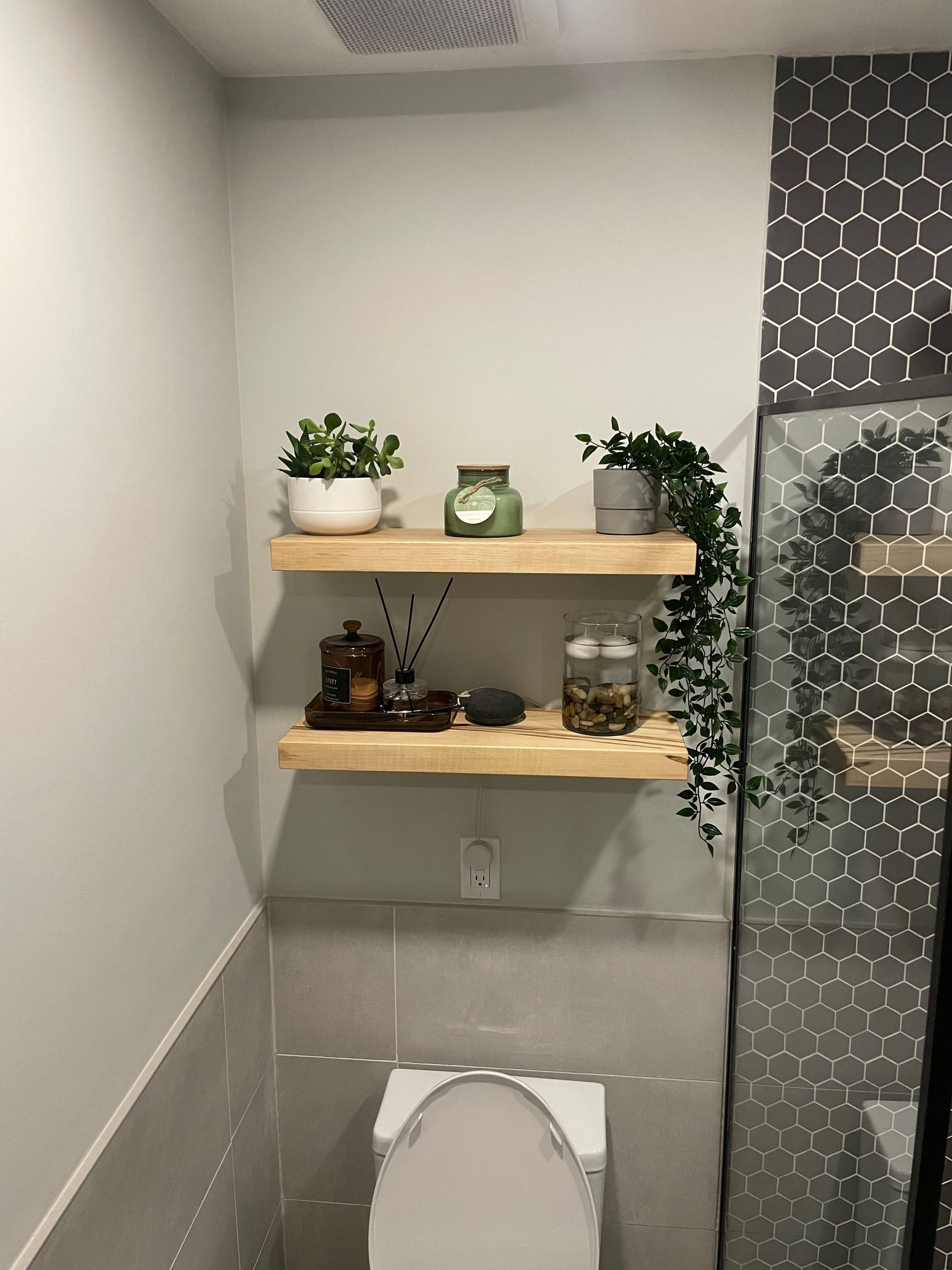 Two wooden shelves with plants and decor above a toilet in a bathroom. Gray tiles on walls.
