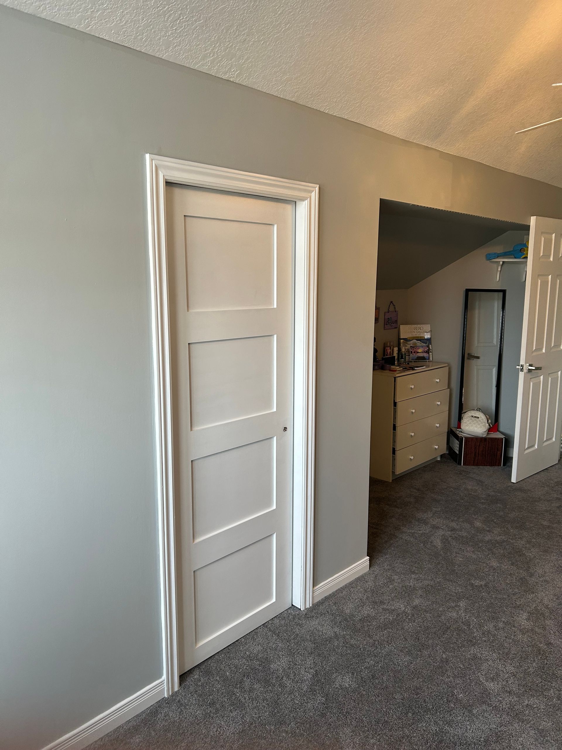 White door with white trim set in gray wall, leading to another room with a chest of drawers.