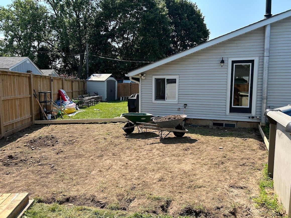 Backyard scene with house, fence, wheelbarrow, and cleared dirt area.