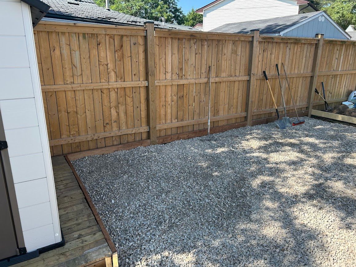 Gravel-covered backyard area next to a wooden fence. Some tools are leaning against the fence.