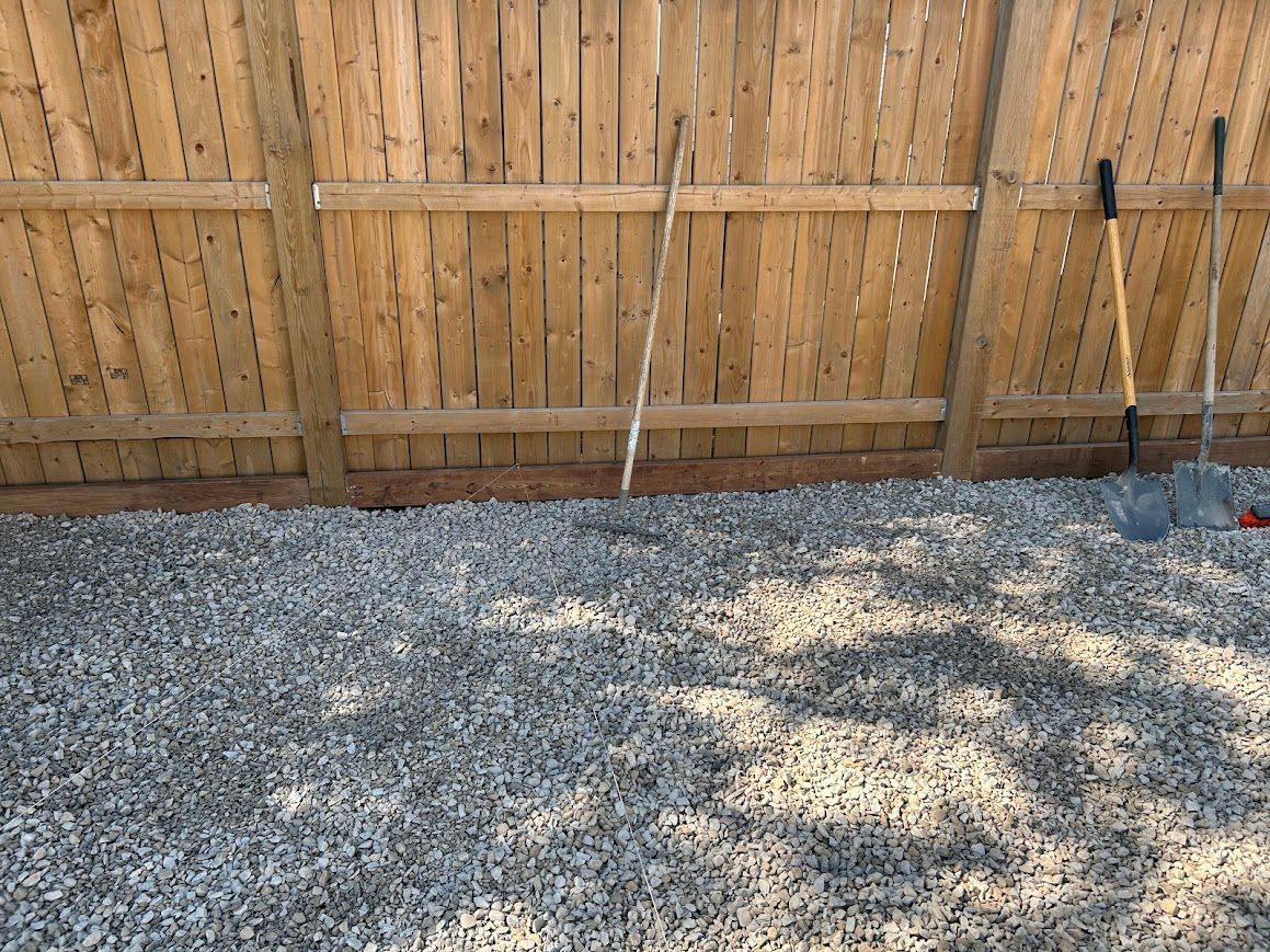 Gravel ground in front of a wooden fence. Gardening tools lean against the fence.