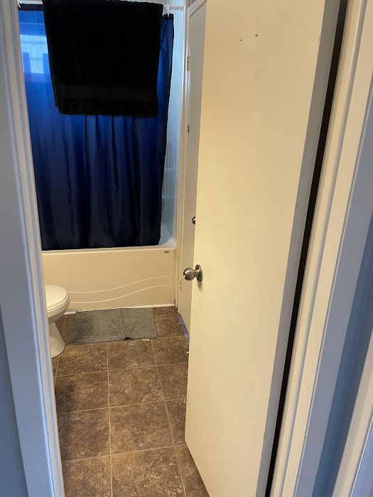 Bathroom interior: white door ajar, showing tub with blue curtain, toilet, brown floor, gray rug, and black towel.