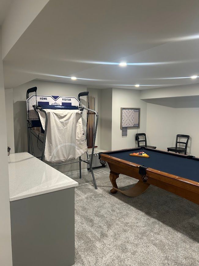 A finished basement recreation room with a pool table, arcade game, and seating. Gray carpet and neutral walls.