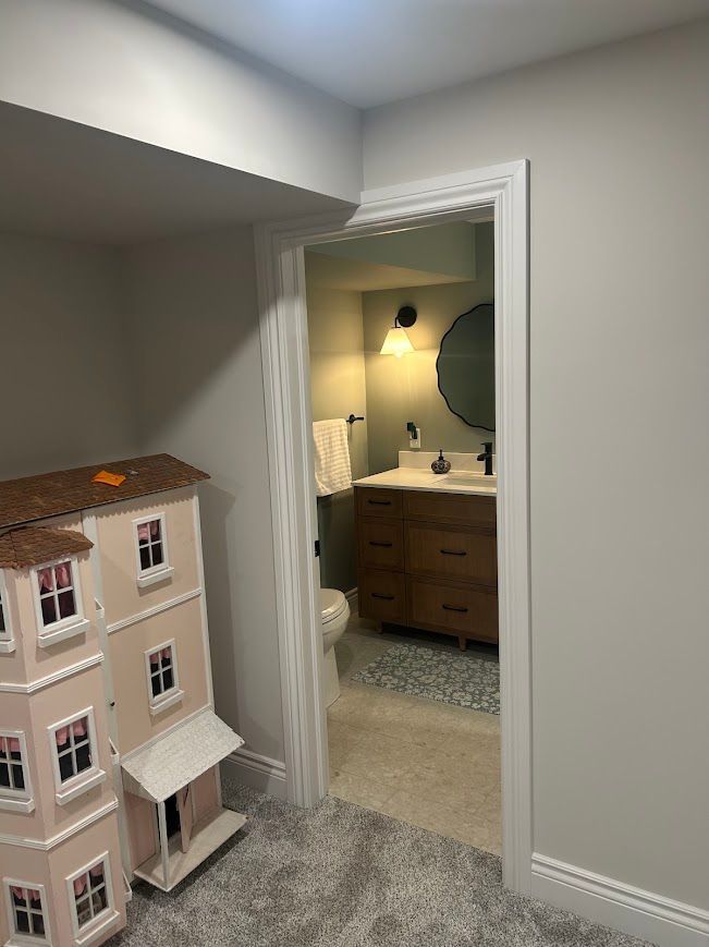 Doorway to a bathroom with a wooden vanity, round mirror, and patterned tile floor; a dollhouse sits nearby.