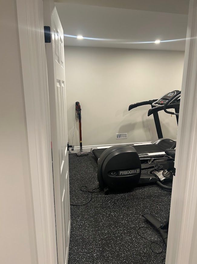 Doorway view of a home gym with treadmill, elliptical, and vacuum cleaner. Black speckled flooring, light gray walls.