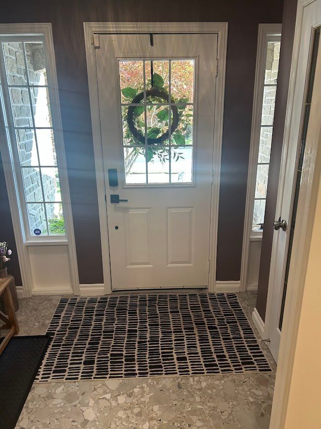 White front door with wreath, flanked by windows. Black and white patterned rug.
