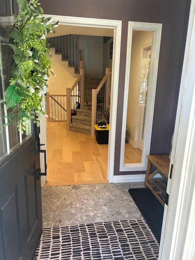 Entryway with open door, showing wood floors, staircase, and a mirror. A wreath hangs on the door.
