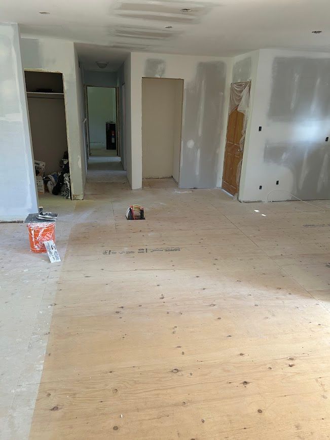 Interior renovation in progress: bare plywood floor, drywall on walls, doorway to hallway, construction materials.