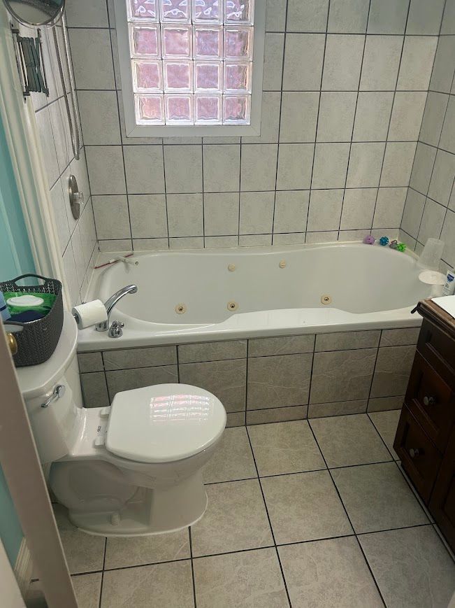 Bathroom with a toilet, a jacuzzi tub, and a window. White tile walls and floor.