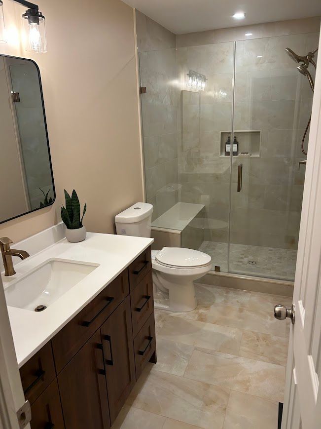 Bathroom with a shower, vanity, and toilet. Light-colored tile on the floor and walls. Brown vanity.