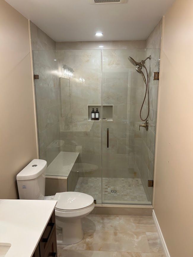 Bathroom with glass shower, neutral tile, toilet, built-in bench, and shower head.