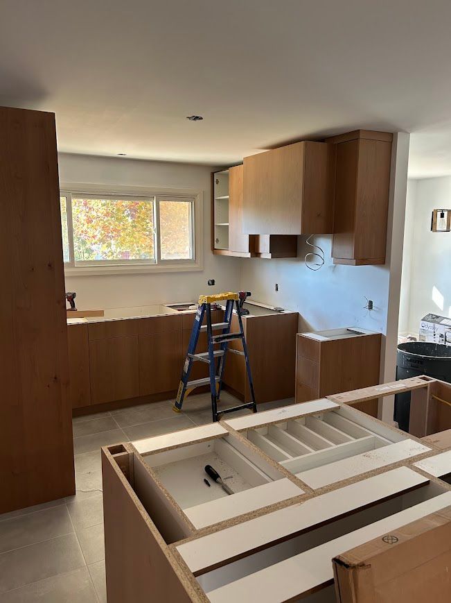Kitchen under construction; wood cabinets and countertop, stepladder, tools visible, unfinished walls, bright light.
