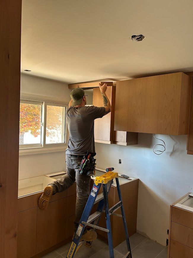 Person installing kitchen cabinets, standing on a ladder. Light brown cabinets and wall with window.