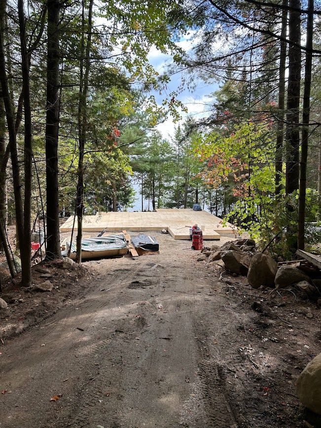 Dirt road leads to a wooden house foundation under construction near a lake, surrounded by trees.