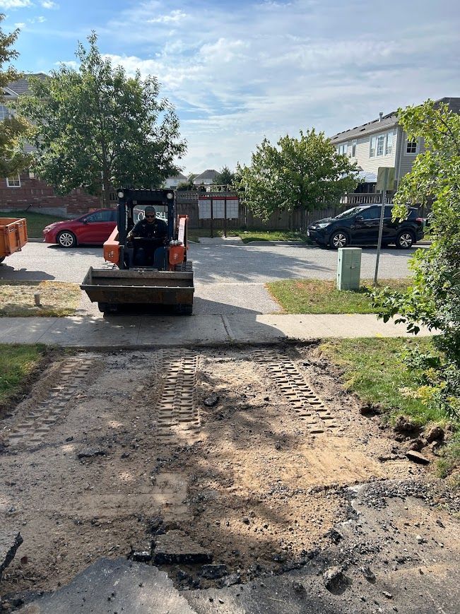 Mini excavator paving a driveway; houses and cars in background.