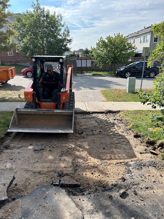 Man operating a small excavator removing asphalt from a sidewalk in front of houses.
