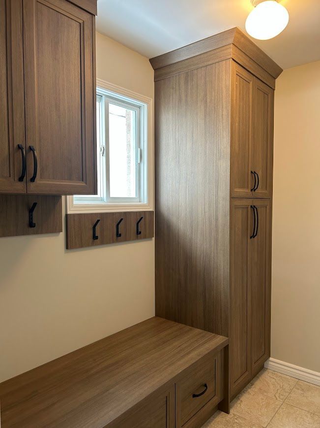 Wooden storage cabinets and bench in a laundry room, with a window and hooks.