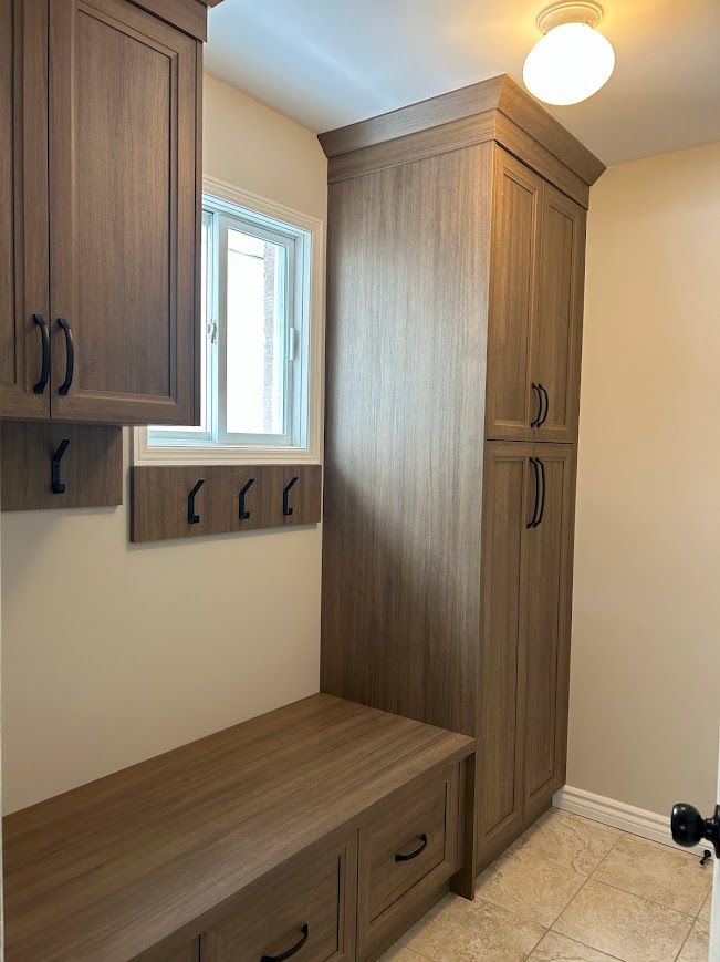 Laundry room with brown cabinets, bench, and hooks. Window, beige walls, and light fixture.