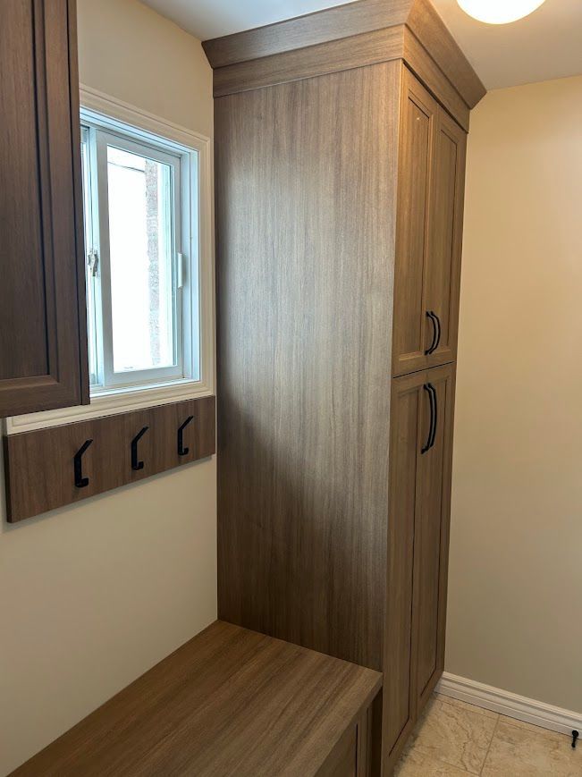 Tall wood storage cabinet beside a window with a coat rack and bench in a room.