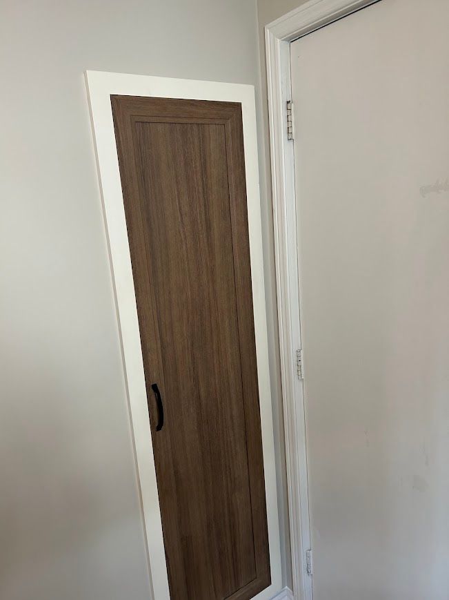 A tall, narrow wooden door with a dark handle and white trim set into a beige wall.