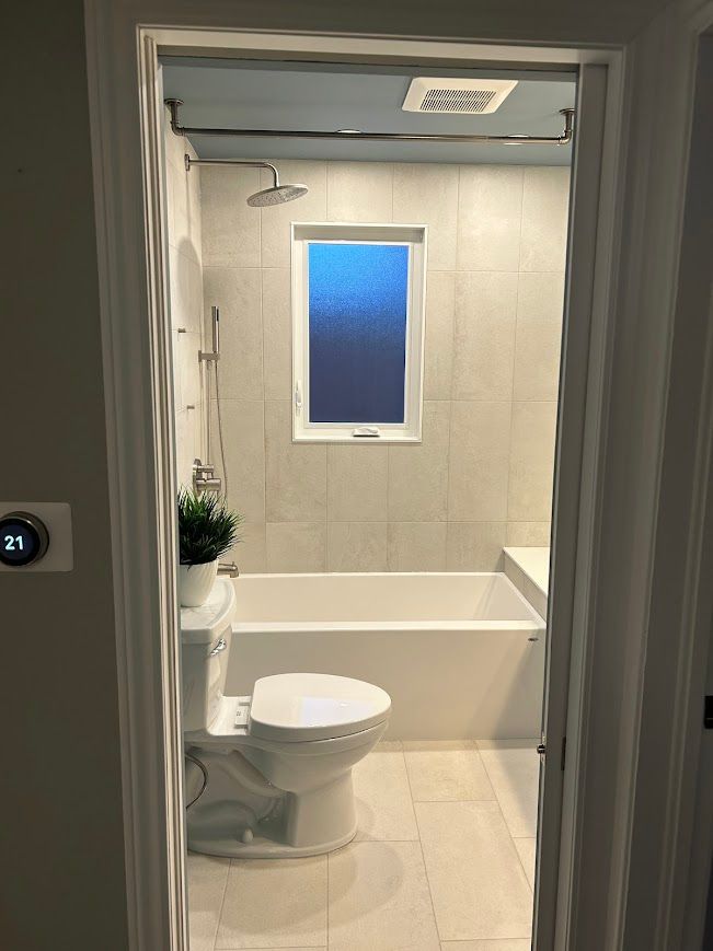 Bathroom with white tub, toilet, and tiled walls. A window is above the tub.