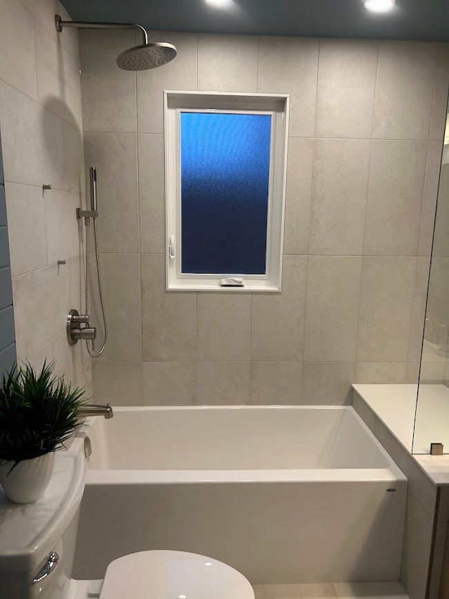 Bathroom with white tub, shower head, window, light gray tile, and toilet.