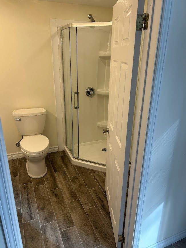 Bathroom with a toilet, shower, and partially open white door. Wood-look flooring and beige walls.