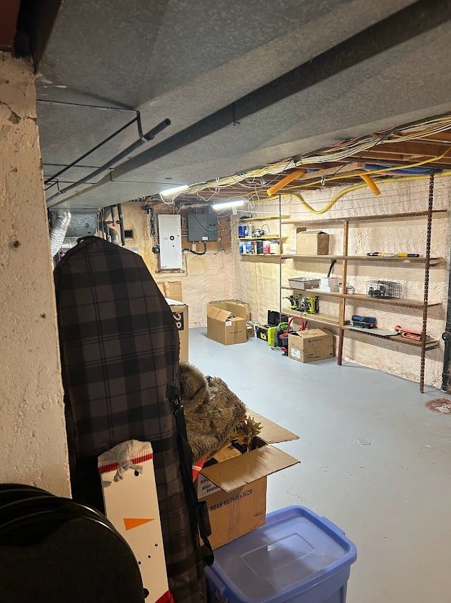 Cluttered basement storage area with shelves, boxes, and various items. Gray floor, concrete ceiling, and limited natural light.