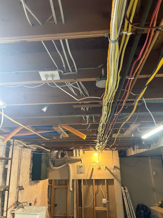 Exposed electrical wiring on a basement ceiling with multiple colored cables and outlets.