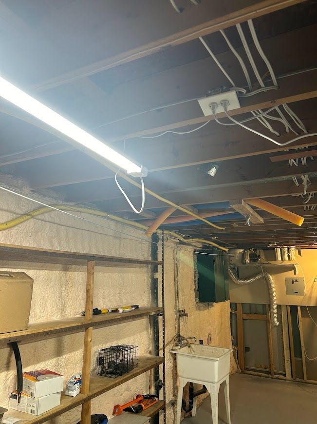 Basement interior with exposed beams, shelving, laundry sink, electrical wiring, and a fluorescent light.