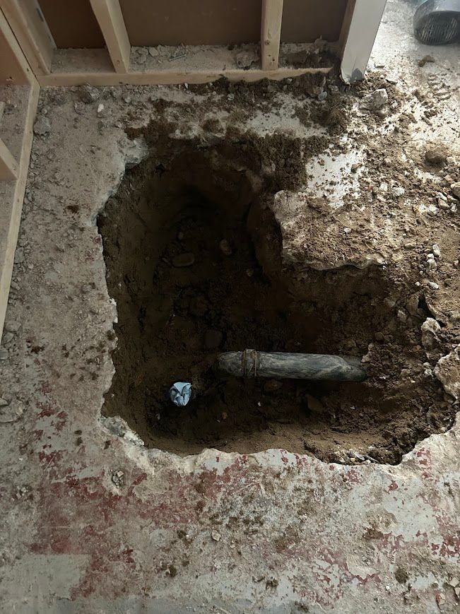 Hole in floor reveals buried pipe. Brown earth surrounds the concrete. Light, wooden frame in the background.