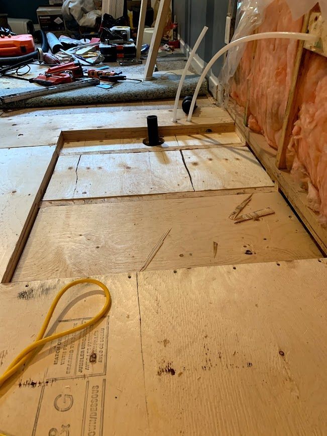 Wooden subfloor with a rectangular opening, exposed pipes, and a partially constructed wall. Construction scene.