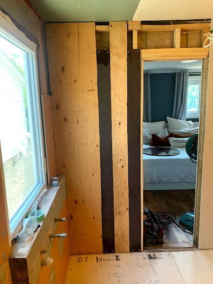Bathroom renovation, partially constructed. Plywood wall with dark vertical beams, doorway to bedroom with bed visible.