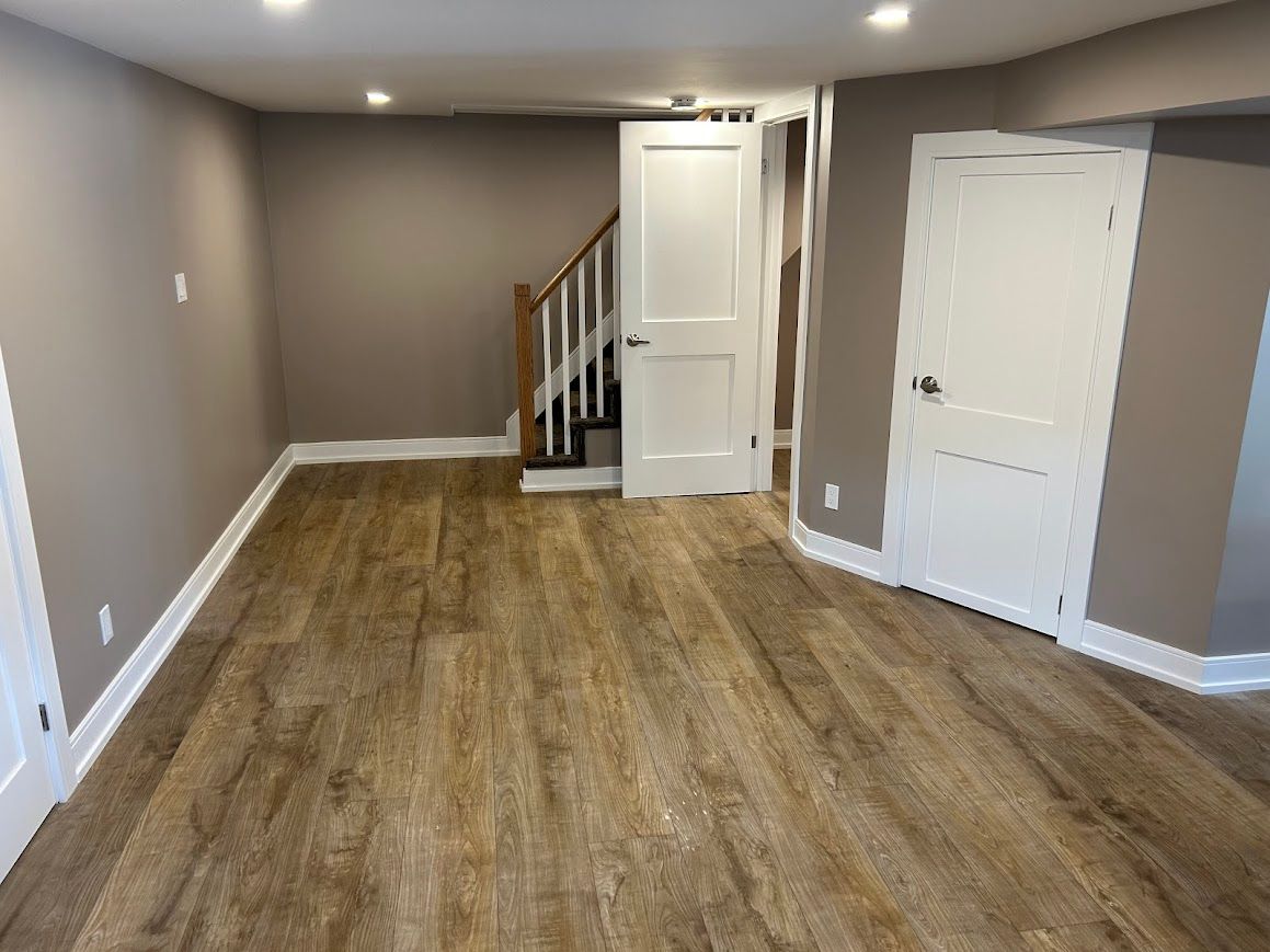 Empty room with wood-look flooring, tan walls, white doors, and staircase.