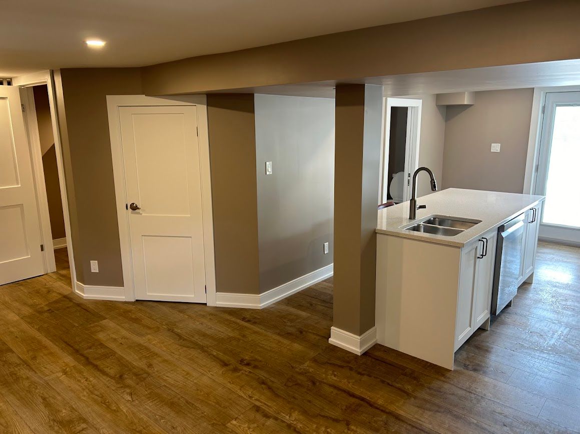 Basement kitchen with white cabinets, island, and wood-look flooring. Tan walls, white trim and door.