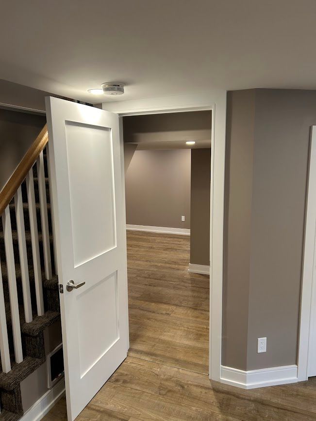 Open white door leading into a room with brown walls and wood-look flooring, beside a staircase.
