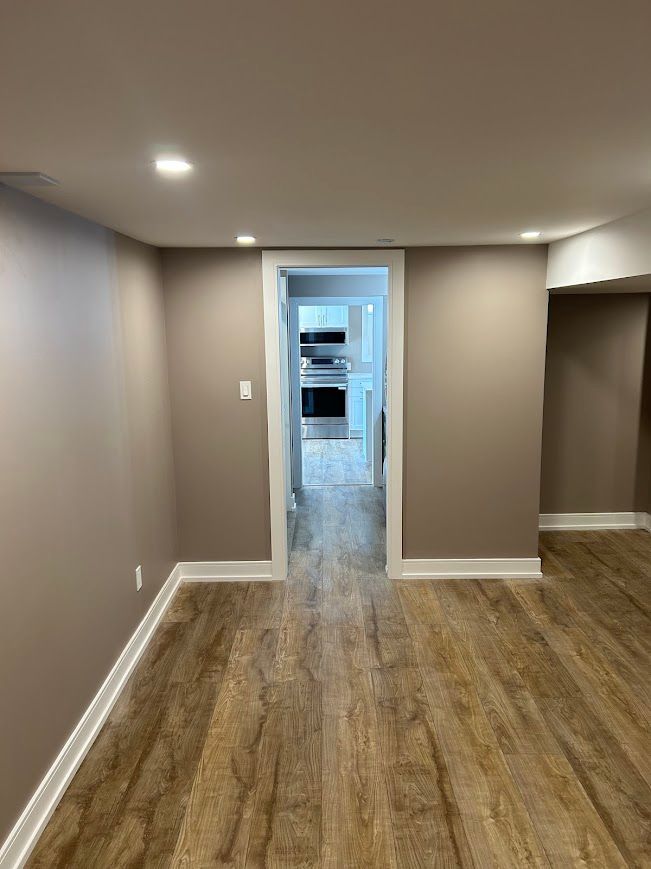 Interior view of a room with wood-look flooring, light brown walls, and an opening to a kitchen.
