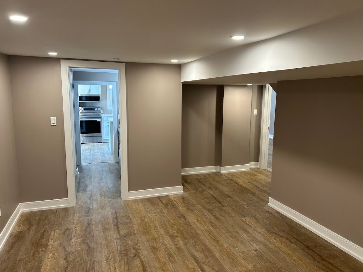 Room with tan walls, white trim, and wood-look flooring. A doorway leads to another room.