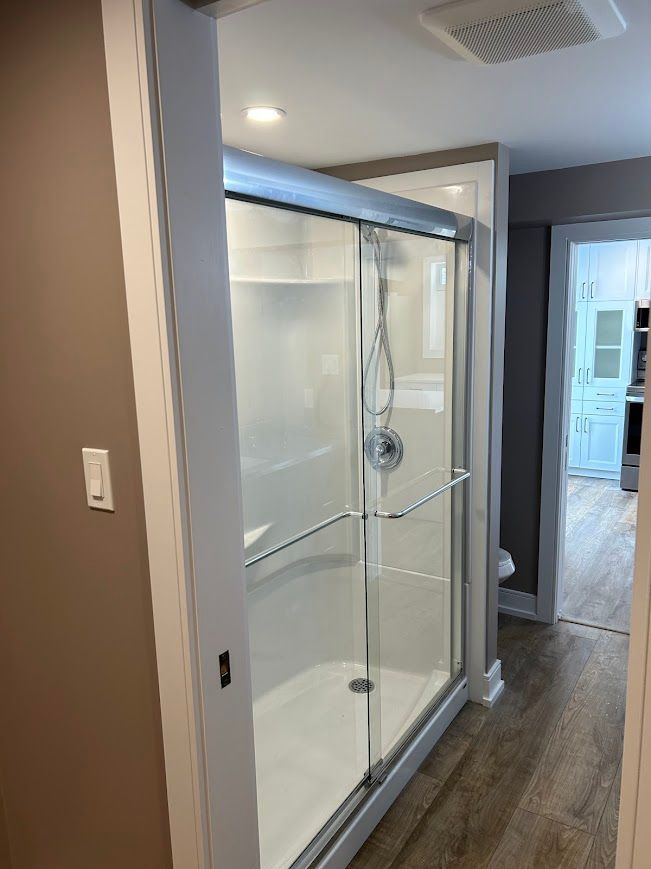 Shower with sliding glass doors in a bathroom hallway. White walls, gray door frame, and light wood flooring.