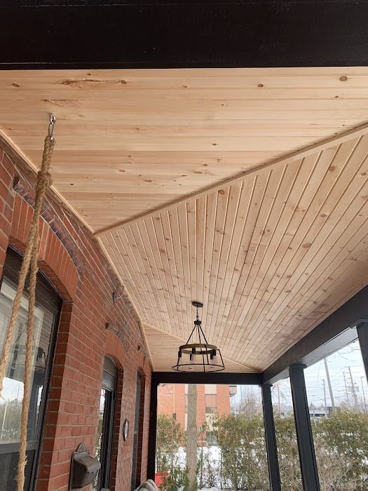 Wood-paneled porch ceiling with a hanging light fixture and a brick wall. Sunlight streams in.