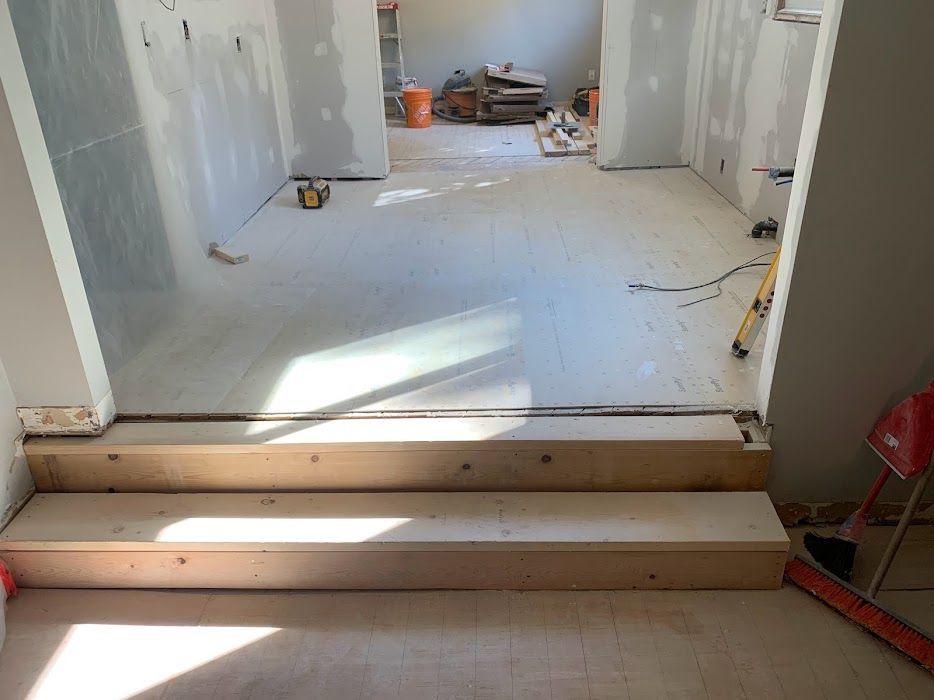 Interior construction site with two wooden steps leading into a room. Drywall and tools visible.