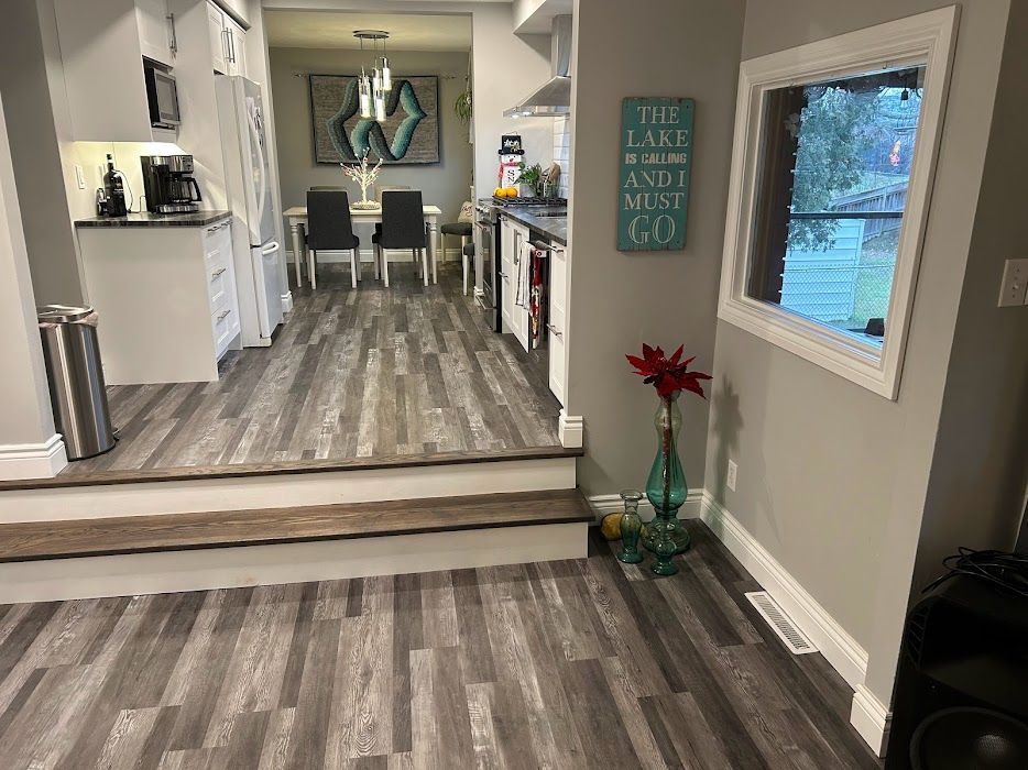 A room with wood-look flooring, leading to a dining area. Steps lead up to the kitchen.