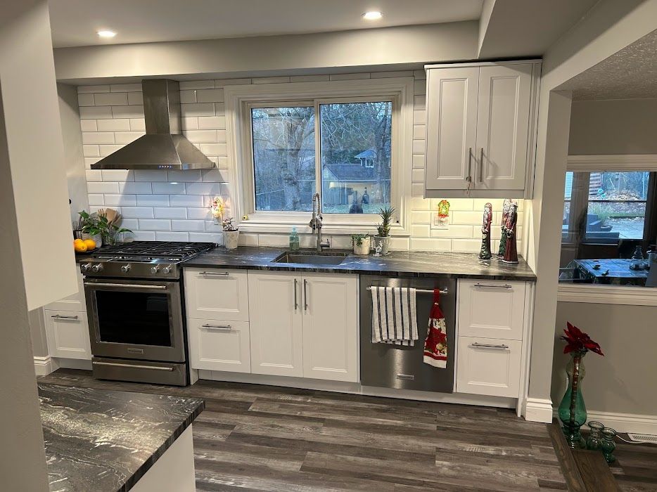 White kitchen with stainless steel appliances, white cabinets, dark countertops, and a window.