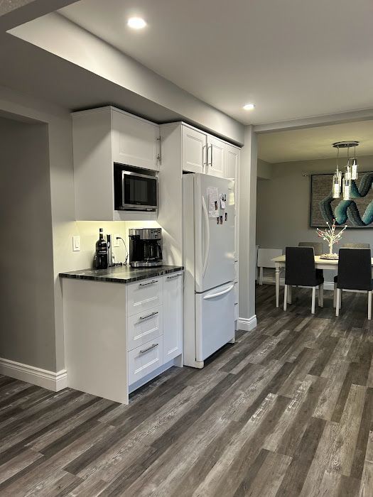 White kitchen cabinets with black countertop, microwave, refrigerator, and coffee maker, open to a dining area with dark chairs.