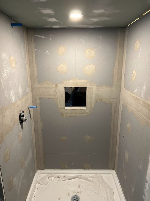 Interior view of a shower stall under construction; gray walls with drywall and a centered square window opening.
