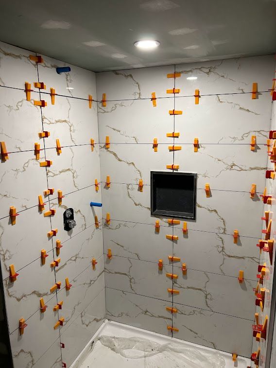 Shower with white marble-look tiles, orange tile leveling clips, and a black recessed shelf.