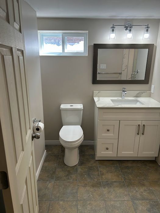 Bathroom with white vanity, toilet, and a window. Beige walls and brown tile floor.