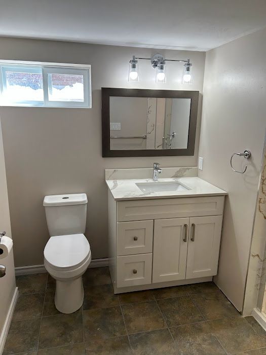 Bathroom with white vanity, toilet, and a window. Light-colored walls and dark tile flooring.