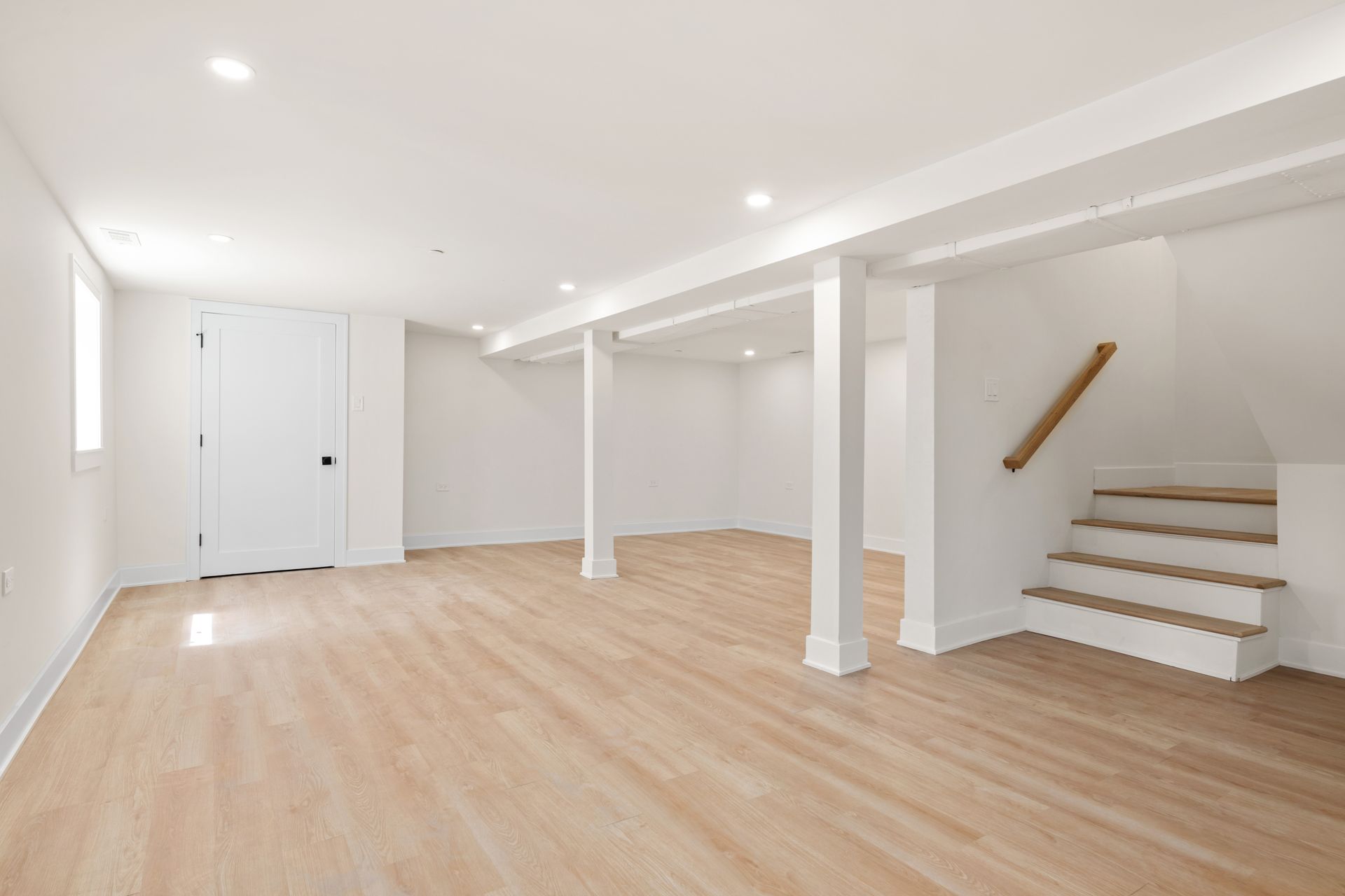 Empty, bright white basement room with wood-look floors, support columns, and a staircase with a wooden handrail.