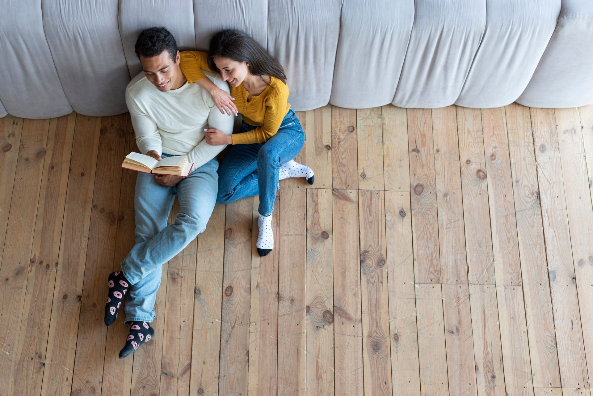 Couple reading a book together on wooden floor near a couch. Woman is leaning on man, both are smiling.